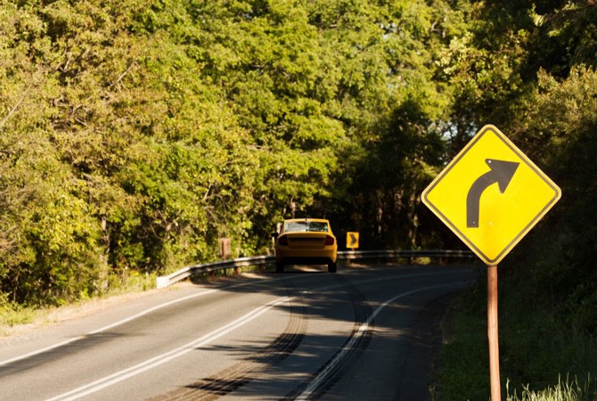 A yellow car, driving through a wooded area, misses a right turn sign and swerves off the road due to steering and suspension parts failure.