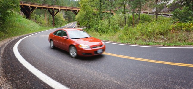 Car driving on an open road around a bend with trees and a small bridge behind